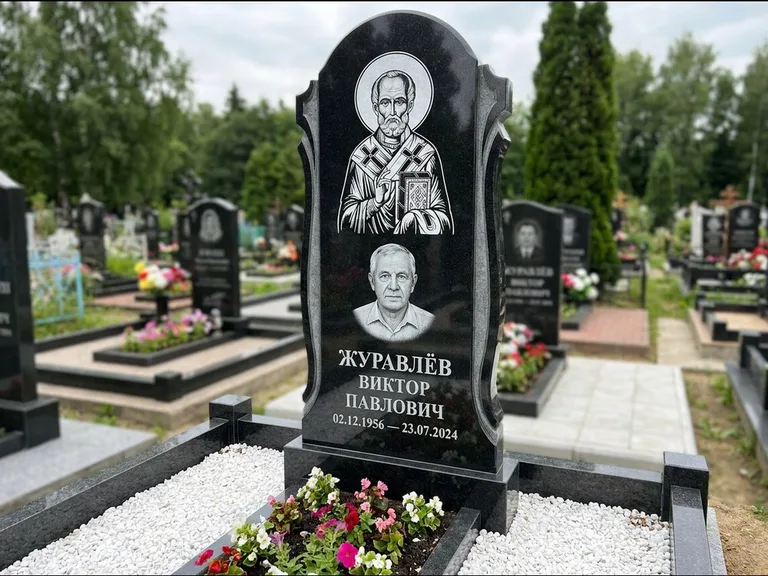 Памятник с гравировкой иконы: стела с образом в полный рост на обороте — гравировка образа Матроны Московской, низкие кусты спиреи в цвету, работа «Лебедева» в Екатеринбурге